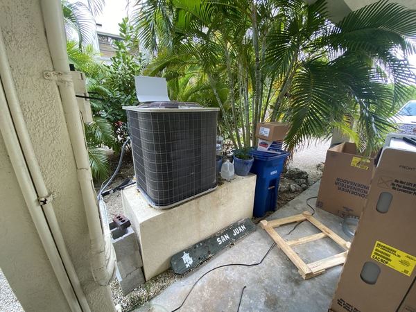 New outdoor AC condenser installed on a raised concrete stand beside a home, surrounded by palms
