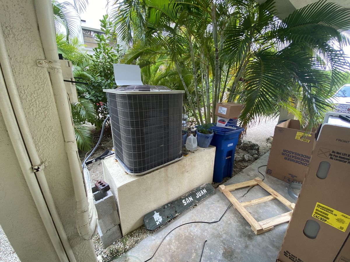 New outdoor AC condenser installed on a raised concrete stand beside a home, surrounded by palms