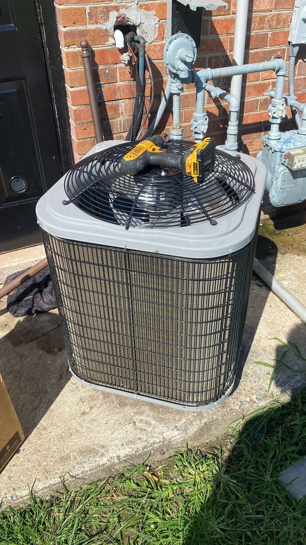 Outdoor AC condenser being serviced beside a brick wall, with a tool resting on the fan grille