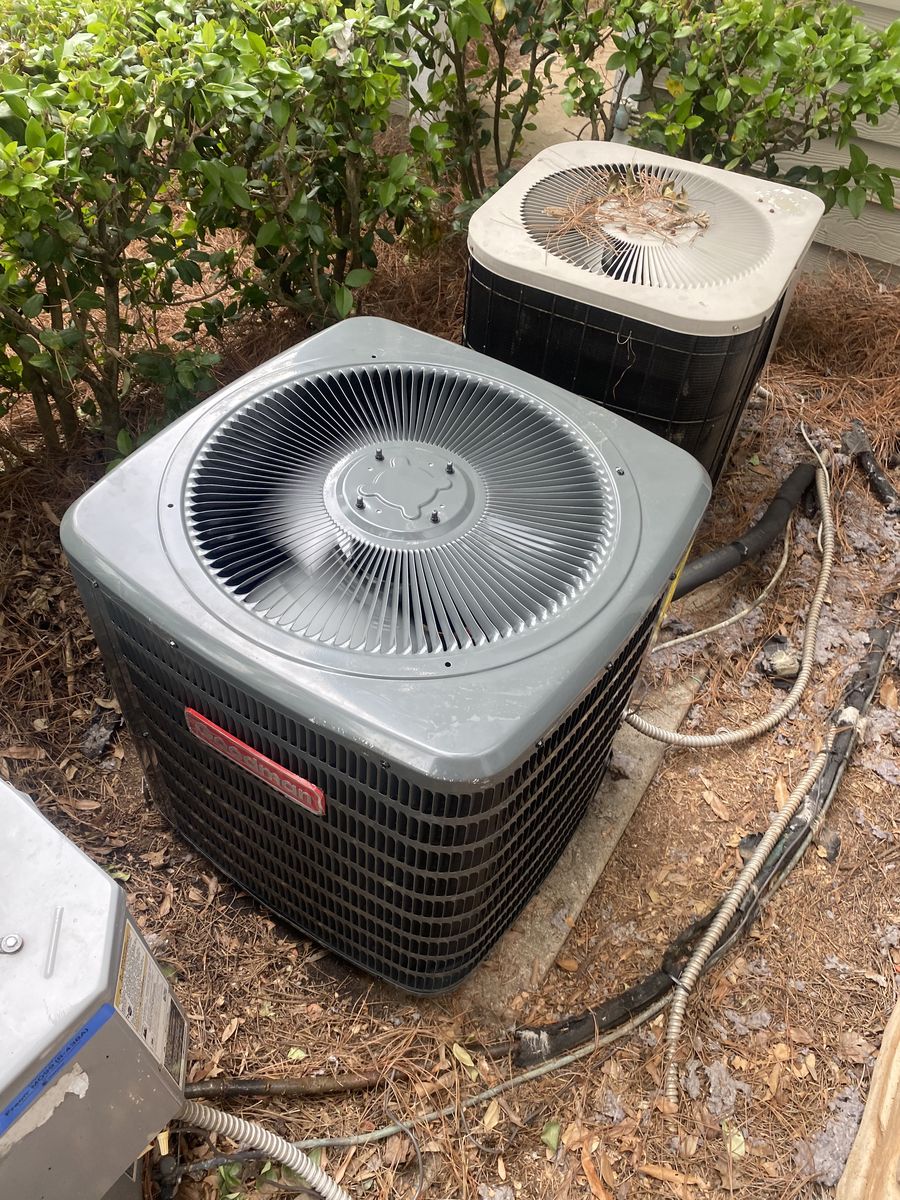Two outdoor AC condensers side by side among shrubs, with refrigerant lines connected